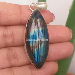 May include: A silver pendant with a large, oval labradorite stone. The stone displays iridescent blue, green, and gold hues. The pendant has a simple silver bail and is held by a hand. The text "@gemssilverjewls" is visible.