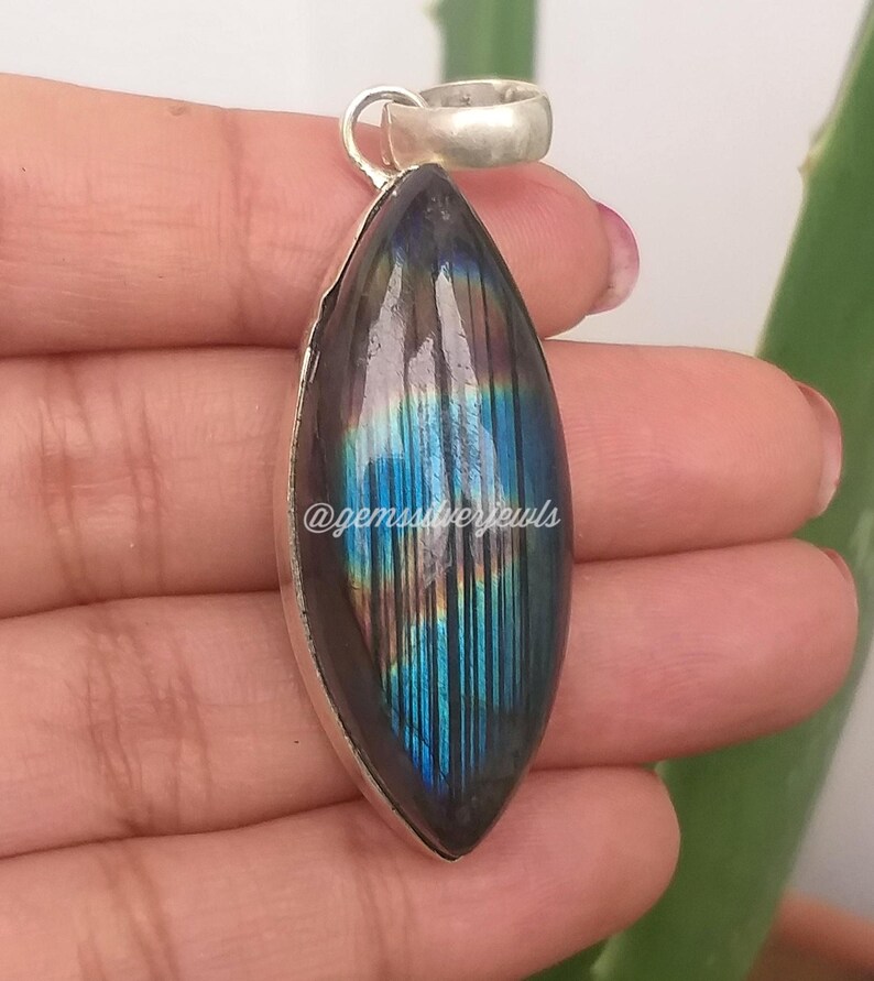 May include: A silver pendant with a large, oval labradorite stone. The stone displays iridescent blue, green, and gold hues. The pendant has a simple silver bail and is held by a hand. The text "@gemssilverjewls" is visible.