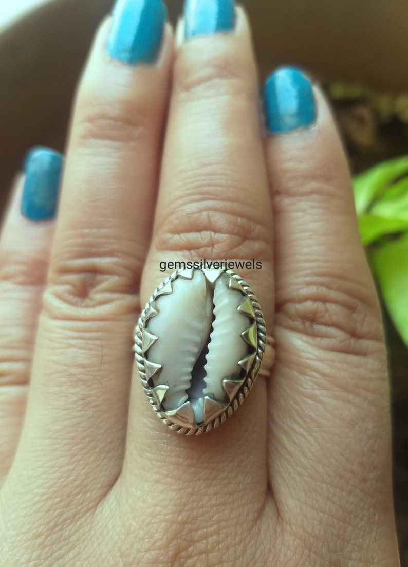 May include: A silver ring with a white cowrie shell set in a silver bezel. The shell is surrounded by a silver rope design and a row of silver triangles.