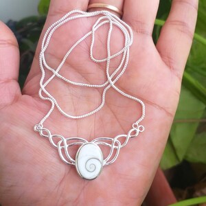 May include: A silver chain necklace with a silver pendant featuring a white and green shell. The pendant is in the shape of an oval with a Celtic knot design.