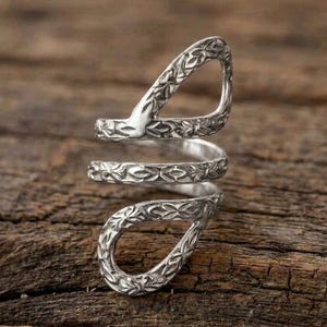 May include: A silver-toned metal ring featuring an open, spiraling design that wraps around the finger. The surface of the metal is textured with a repeating, intricate leaf-like pattern throughout the entire length of the band.