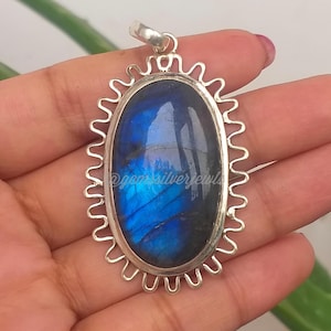 Op de afbeelding: Een zilveren hanger met een ovale, iriserende blauwe edelsteen. De edelsteen is gezet in een decoratief, geschulpt zilveren frame. De hanger heeft een kleine lus aan de bovenkant voor een ketting. De tekst "@gemsilverjewels" is zichtbaar.