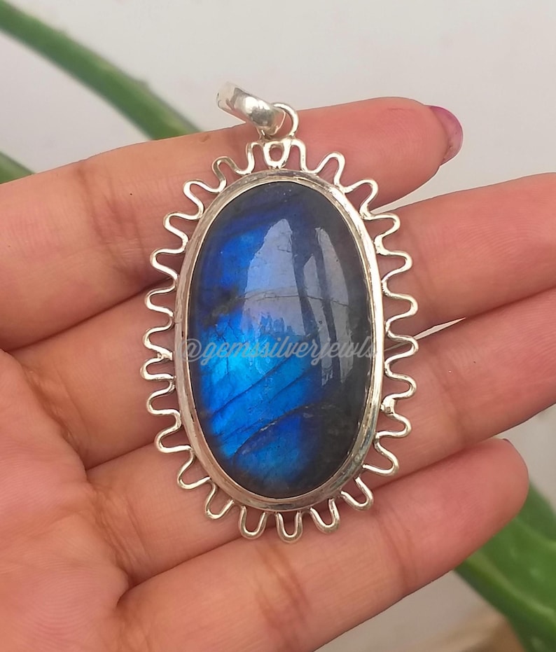 Op de afbeelding: Een zilveren hanger met een ovale, iriserende blauwe edelsteen. De edelsteen is gezet in een decoratief, geschulpt zilveren frame. De hanger heeft een kleine lus aan de bovenkant voor een ketting. De tekst "@gemsilverjewels" is zichtbaar.