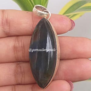 May include: A dark blue and silver pendant with a teardrop-shaped gemstone. The gemstone has a polished surface and is set in a silver bezel. The pendant has a silver loop at the top for a chain. The text "@gemssilverjewls" is visible.