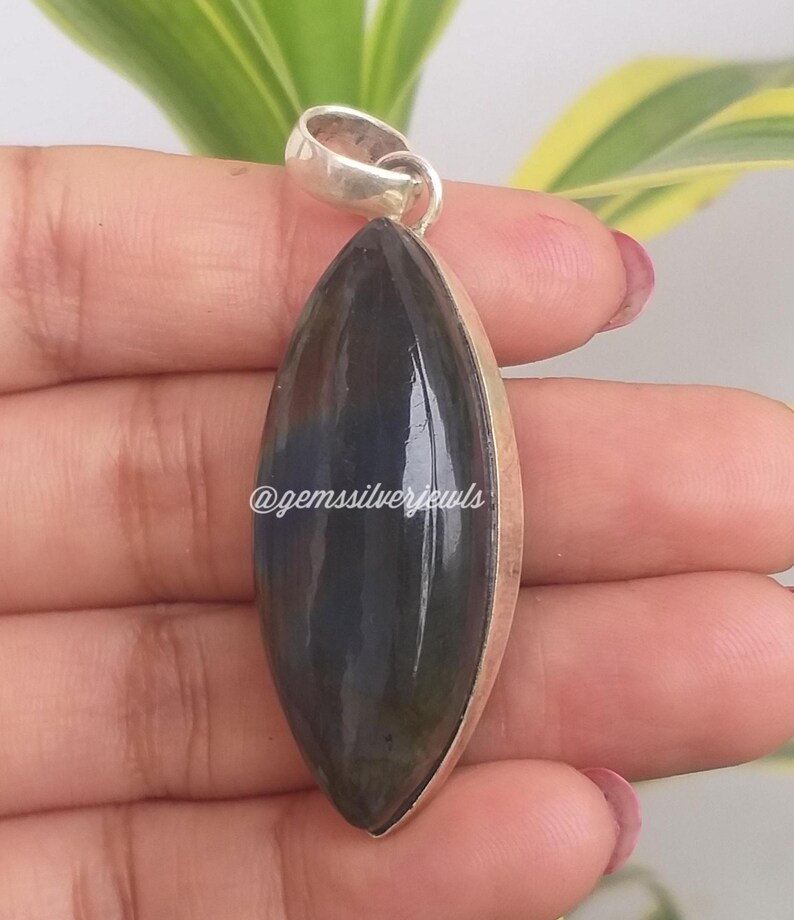 May include: A dark blue and silver pendant with a teardrop-shaped gemstone. The gemstone has a polished surface and is set in a silver bezel. The pendant has a silver loop at the top for a chain. The text "@gemssilverjewls" is visible.