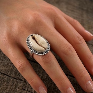 Peut inclure: Une bague en argent ornée d'un coquillage cauri ovale en son centre. Le coquillage est entouré d'une bordure décorative composée de petites perles d'argent et d'un détail en fil d'argent torsadé. L'anneau est en argent lisse et poli.