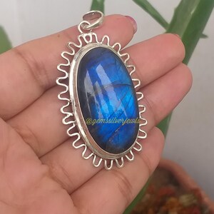 Op de afbeelding: Ovale labradoriet hanger met een zilverkleurig, golvend frame. De blauwe edelsteen heeft interne patronen en is gezet in een zilveren bezel. De hanger is opgehangen aan een zilveren lus. De tekst "@gemssilverjewls" is zichtbaar.