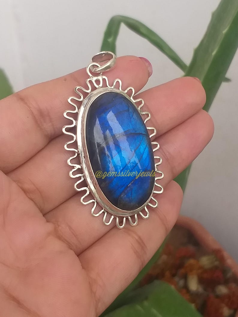 Op de afbeelding: Ovale labradoriet hanger met een zilverkleurig, golvend frame. De blauwe edelsteen heeft interne patronen en is gezet in een zilveren bezel. De hanger is opgehangen aan een zilveren lus. De tekst "@gemssilverjewls" is zichtbaar.