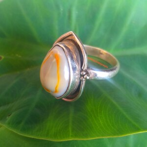 K&ouml;nnte beinhalten: Ein silberner Ring mit einem tropfenf&ouml;rmigen Achat-Stein. Der Stein ist wei&szlig; mit orangefarbenen und braunen Wirbeln. Der Ring liegt auf einem gr&uuml;nen Blatt.