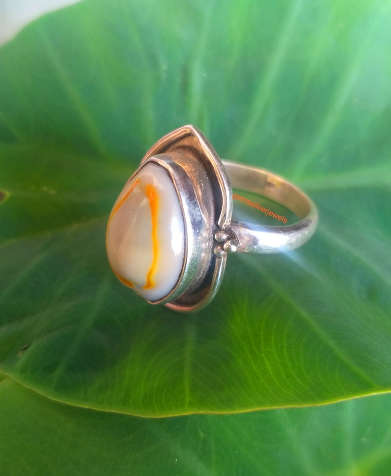 K&ouml;nnte beinhalten: Ein silberner Ring mit einem tropfenf&ouml;rmigen Achat-Stein. Der Stein ist wei&szlig; mit orangefarbenen und braunen Wirbeln. Der Ring liegt auf einem gr&uuml;nen Blatt.