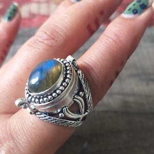 May include: A silver ring featuring an oval-shaped labradorite gemstone with blue and gold iridescence. The stone is set in a beaded bezel, and the band displays intricate metalwork with textured, braided-style patterns and openwork details.