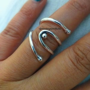 May include: A silver-colored ring wraps around a finger, featuring a spiral design with a small ball accent. The ring's smooth, polished surface reflects light, creating a modern and elegant look. The ring is a piece of jewelry.