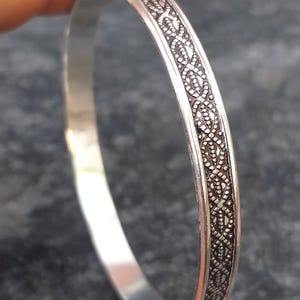 May include: A silver bangle bracelet with an intricate, repeating pattern around the band. The bracelet is smooth and polished, with a detailed design in a darker tone. The bracelet is held by a hand, showcasing its design.