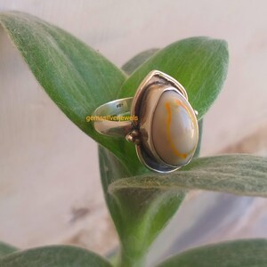 K&ouml;nnte beinhalten: Ein silberner Ring mit einem wei&szlig;en und orangefarbenen Achat-Stein. Der Ring ist in einer silbernen Fassung mit einem dekorativen Design gefasst. Der Ring liegt auf einem gr&uuml;nen Blatt.