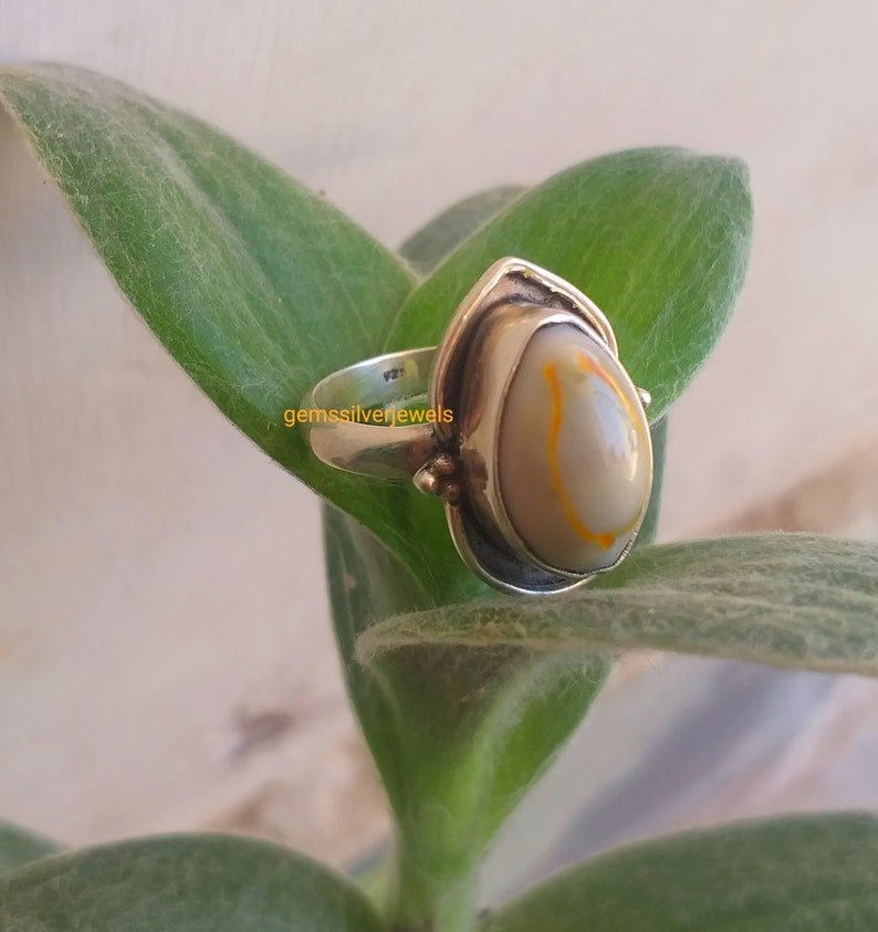 K&ouml;nnte beinhalten: Ein silberner Ring mit einem wei&szlig;en und orangefarbenen Achat-Stein. Der Ring ist in einer silbernen Fassung mit einem dekorativen Design gefasst. Der Ring liegt auf einem gr&uuml;nen Blatt.