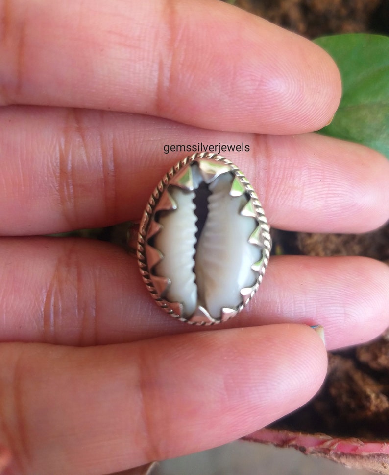 May include: A silver ring with a white cowrie shell set in the center. The shell is surrounded by a silver border with a decorative design.