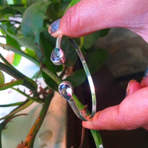 May include: A silver bangle bracelet with two teardrop-shaped iridescent gemstones. The bracelet is open and has a simple design.