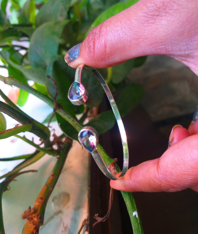 May include: A silver bangle bracelet with two teardrop-shaped iridescent gemstones. The bracelet is open and has a simple design.