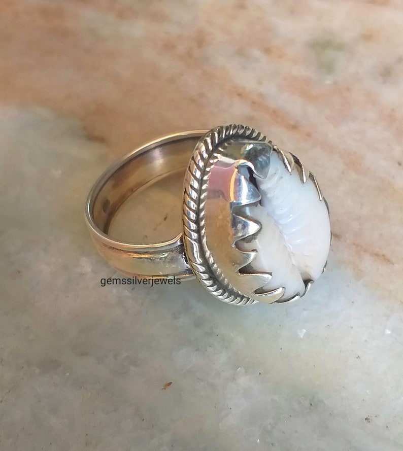 May include: A silver ring with a white shell set in a silver bezel. The ring has a rope-like design around the bezel.