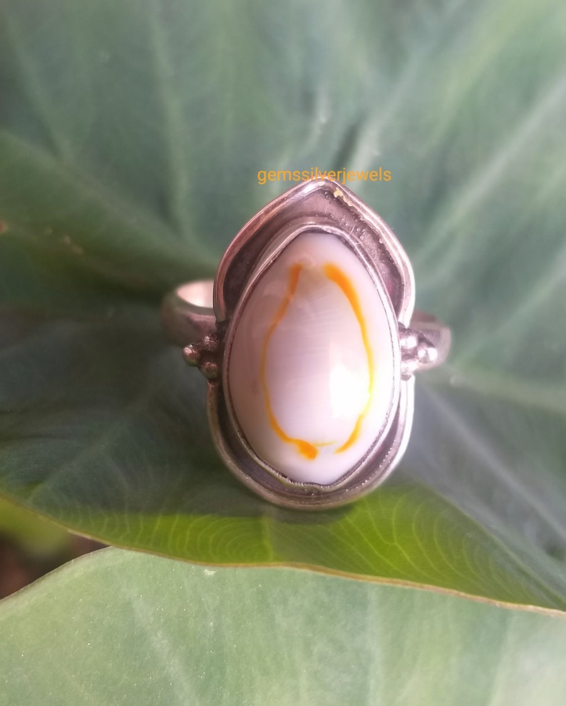 K&ouml;nnte beinhalten: Ein silberner Ring mit einer wei&szlig;en und orangefarbenen Muscheleinlage. Der Ring hat eine Tropfenform und ist in eine silberne Band gefasst.