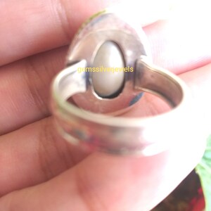 May include: A silver ring with a white oval gemstone set in a bezel setting. The ring has a wide band with a smooth finish. The text "gemssilverjewels" is engraved on the back of the ring.