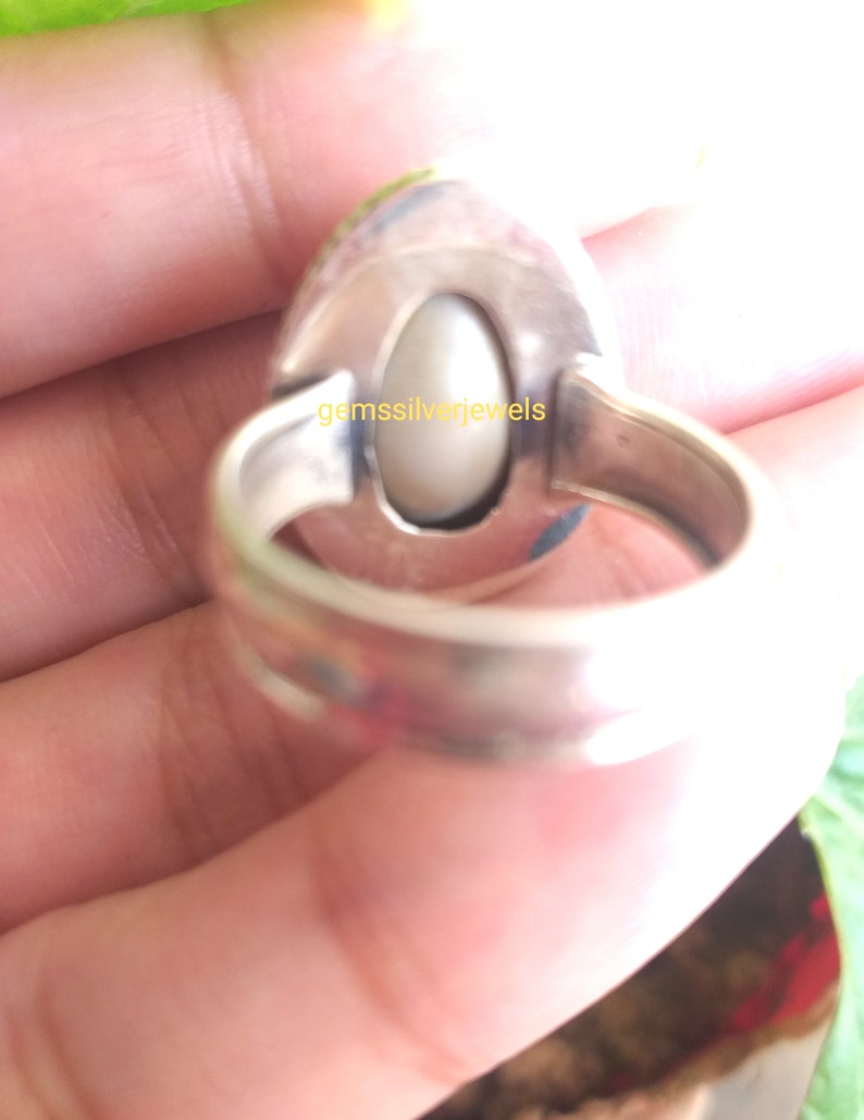 May include: A silver ring with a white oval gemstone set in a bezel setting. The ring has a wide band with a smooth finish. The text "gemssilverjewels" is engraved on the back of the ring.