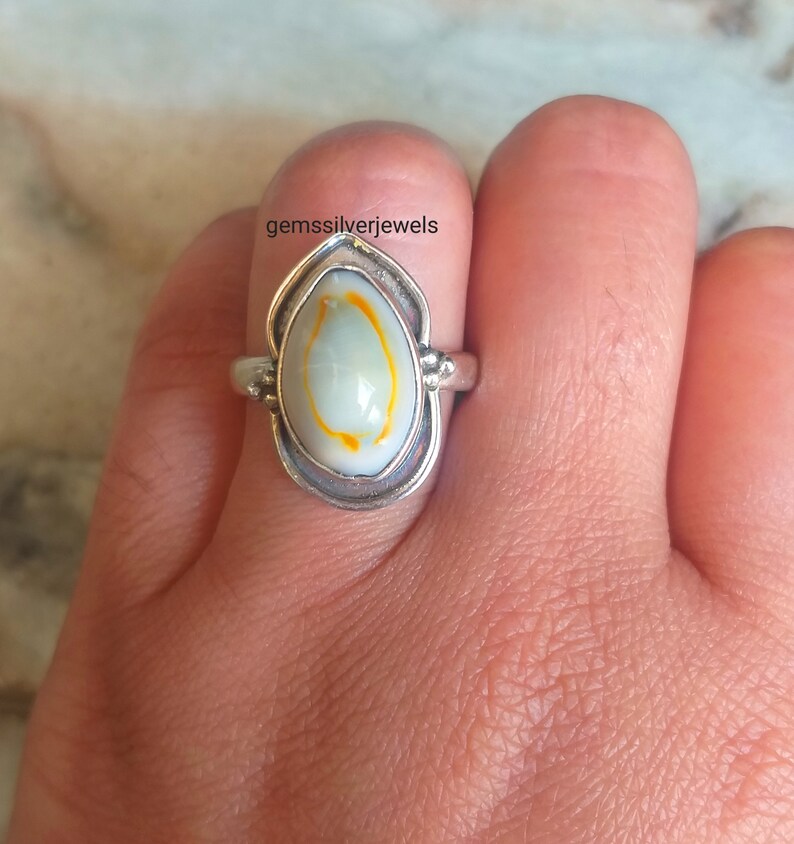 K&ouml;nnte beinhalten: Ein silberner Ring mit einer wei&szlig;en und gelben Muschel, die in einer silbernen Fassung sitzt. Der Ring wird an einem Finger getragen.