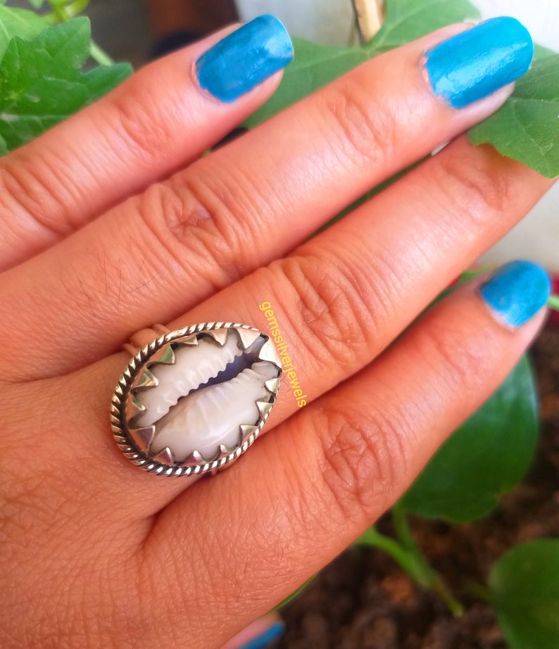May include: A silver ring with a white cowrie shell set in the center. The ring is worn on a hand with blue nail polish.