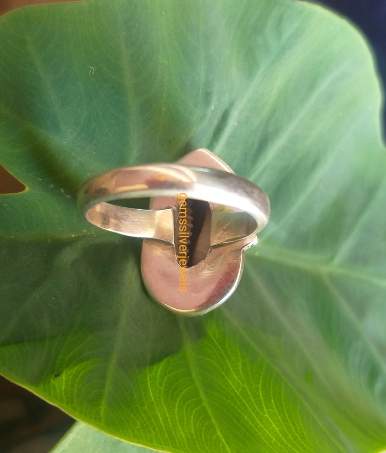 K&ouml;nnte beinhalten: Ein silberner Ring mit einem dunkelgr&uuml;nen Edelstein, der in eine silberne Fassung gefasst ist. Der Ring liegt auf einem gr&uuml;nen Blatt.