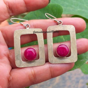 Puede incluir: Pendientes de plata con marco rectangular y acabado texturizado. Cada pendiente presenta una vibrante gema redonda de color magenta. Los pendientes se sujetan con ganchos de plata. El texto "GemssilverjewelsMadhua" es visible.
