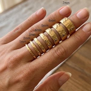 Puede incluir: Un conjunto de siete anillos de color dorado con textura martillada, dispuestos en orden creciente de anchura de 1mm a 7mm. Cada anillo está etiquetado con su medida de anchura correspondiente en milímetros, mostrando la variación del grosor.