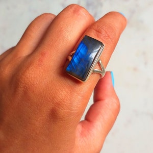 Puede incluir: Un anillo de plata con una piedra rectangular de labradorita azul. El anillo está en un dedo.