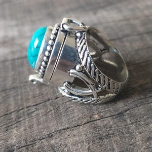 May include: A silver-toned metal ring featuring a prominent, oval-shaped turquoise stone with dark veining. The band displays intricate, raised decorative patterns and a beaded border surrounding the central stone setting, with a hollowed-out design.