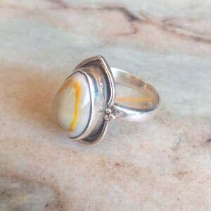 K&ouml;nnte beinhalten: Ein silberner Ring mit einem ovalen wei&szlig;en und orangefarbenen Edelstein, der in einer silbernen Fassung sitzt. Der Ring hat ein schlichtes Band und eine strukturierte Fassung.