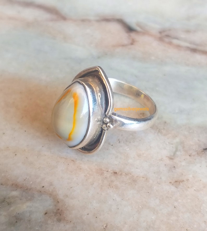 K&ouml;nnte beinhalten: Ein silberner Ring mit einem ovalen wei&szlig;en und orangefarbenen Edelstein, der in einer silbernen Fassung sitzt. Der Ring hat ein schlichtes Band und eine strukturierte Fassung.