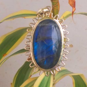 Op de afbeelding: Ovale labradoriet hanger met een goudkleurig, zonvormig frame. De iriserende blauwe steen is geplaatst tegen een achtergrond van groene en gele bladeren. De tekst "@gemssilverjewels" is zichtbaar op de hanger.