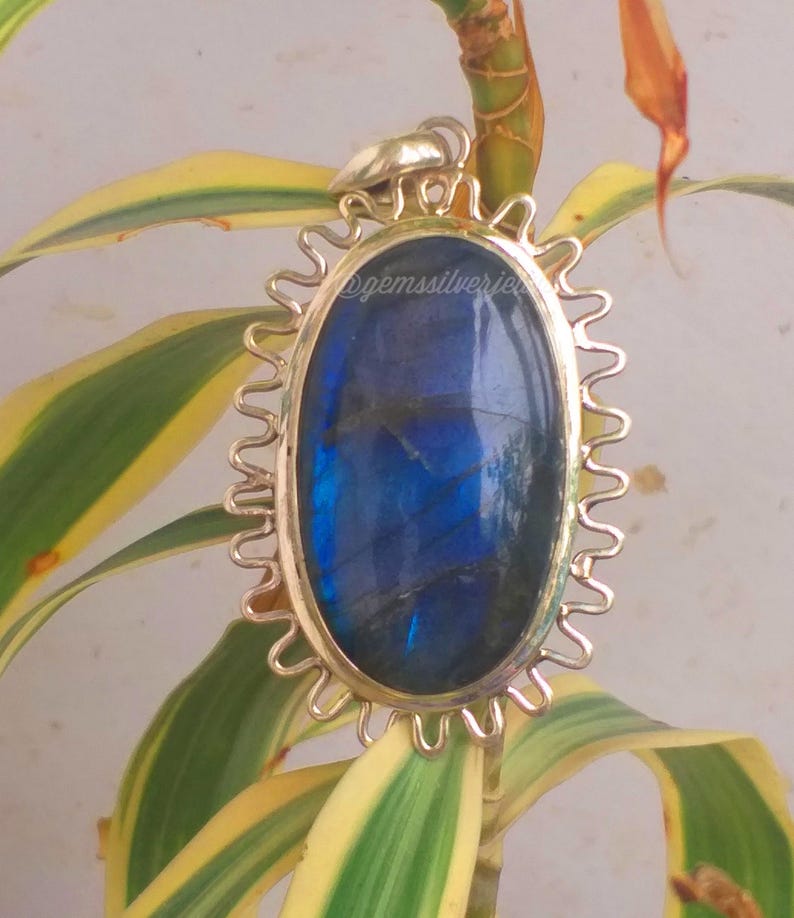 Op de afbeelding: Ovale labradoriet hanger met een goudkleurig, zonvormig frame. De iriserende blauwe steen is geplaatst tegen een achtergrond van groene en gele bladeren. De tekst "@gemssilverjewels" is zichtbaar op de hanger.