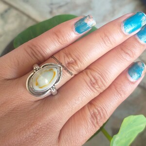 K&ouml;nnte beinhalten: Ein silberner Ring mit einem ovalen Muschelkamee in Wei&szlig; und Gelb. Der Ring wird an einem Finger getragen. Der Text "gemssilverjewels" ist im Bild sichtbar.