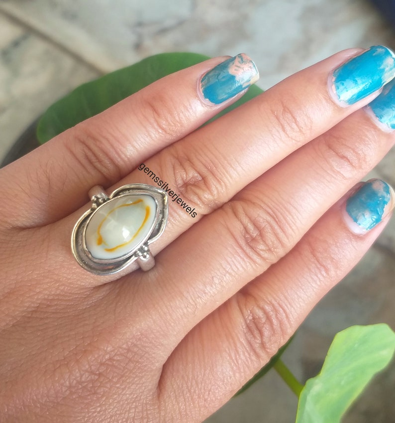 K&ouml;nnte beinhalten: Ein silberner Ring mit einem ovalen Muschelkamee in Wei&szlig; und Gelb. Der Ring wird an einem Finger getragen. Der Text "gemssilverjewels" ist im Bild sichtbar.