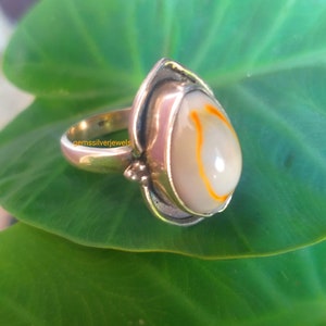 K&ouml;nnte beinhalten: Ein silberner Ring mit einem wei&szlig;en und orangefarbenen Muschelstein, der in Tropfenform gefasst ist. Der Ring liegt auf einem gr&uuml;nen Blatt.