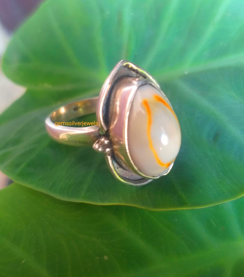 K&ouml;nnte beinhalten: Ein silberner Ring mit einem wei&szlig;en und orangefarbenen Muschelstein, der in Tropfenform gefasst ist. Der Ring liegt auf einem gr&uuml;nen Blatt.