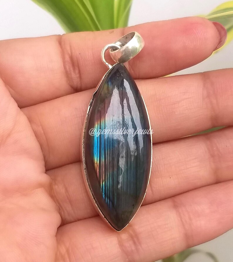 May include: A marquise-shaped pendant with a polished, iridescent labradorite stone set in a silver bezel. The stone displays flashes of blue, green, and gold. The pendant has a silver loop for a chain.