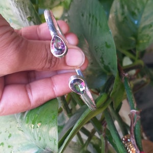 May include: A silver bangle bracelet with two teardrop-shaped iridescent gemstones. The gemstones have a rainbow of colors, including pink, purple, green, and blue.