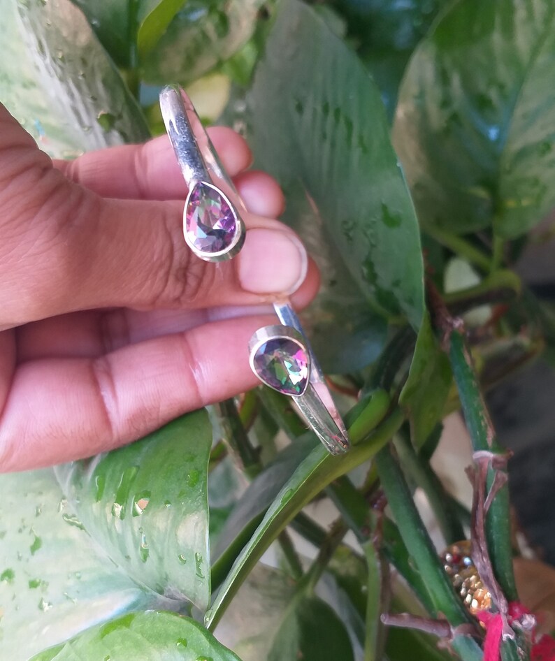 May include: A silver bangle bracelet with two teardrop-shaped iridescent gemstones. The gemstones have a rainbow of colors, including pink, purple, green, and blue.