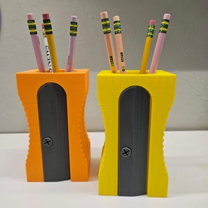 Könnte beinhalten: Zwei 3D-gedruckte Bleistifthalter mit integrierten Spitzer. Einer ist orange und der andere gelb. Beide haben einen schwarzen Spitzer und halten Bleistifte.