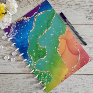 May include: A vibrant notebook featuring a rainbow design with gold glitter detailing. The notebook is open, showing white spiral binding. A black pen and several white disc-shaped objects are placed nearby. A small sprig of flowers is in the upper left corner.