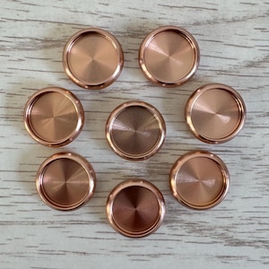May include: Eight copper-coloured, disc-shaped objects with a brushed metal finish are arranged in a circular pattern. Each disc has a slightly raised rim and a central indentation. The discs are on a light wood-grain surface.