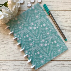 May include: A teal blue notebook with a white floral pattern and white disc binding. The notebook is lying on a white wooden surface with a teal blue pen and white plastic discs.