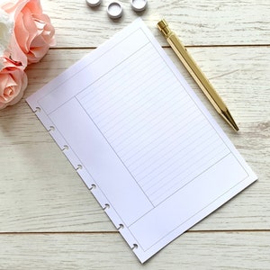 May include: A white lined notebook with a gold pen on a white wooden surface. The notebook has a hole punch on the left side and is open to a blank page. There are five white round stickers on the surface.
