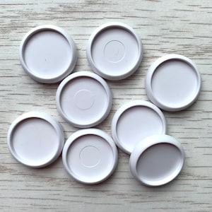 May include: Eight white plastic round buttons with a raised centre circle. The buttons are arranged in a loose cluster on a white wooden surface.
